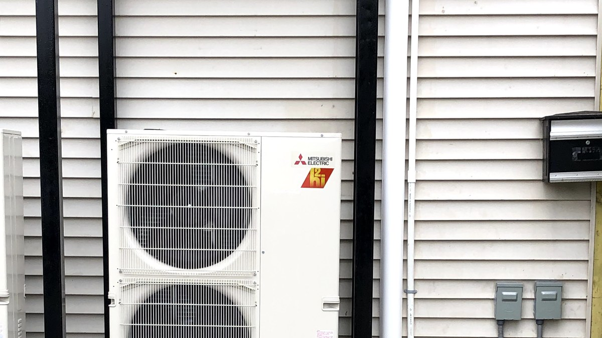 Outdoor mini-split heat pump unit mounted beside a building with visible piping and electrical service access.